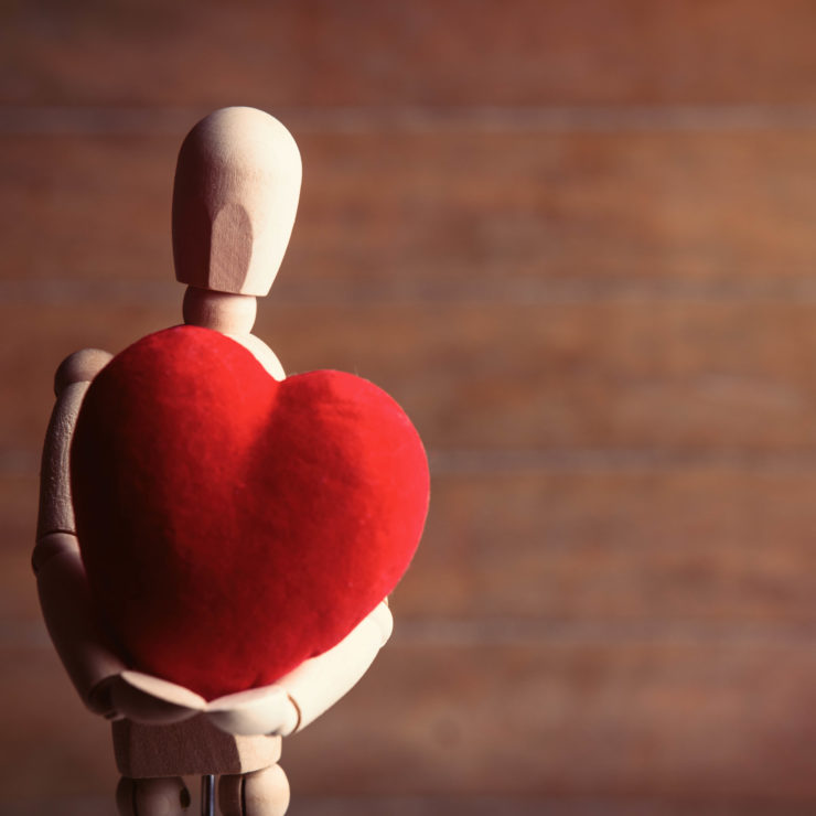 Wood stick figure holding a knitted red heart demonstrating the need for people to practice self-compassion.