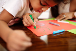Child watching as an adult uses a green crayon to draw. This image demonstrates how adults can teach children well-being.
